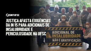 Descrição da imagem: À direita, fotografia de um grupo de TAEs usando equipamentos de proteção, como toucas, máscaras e aventais. Na frente, seguram uma faixa preta e amarela com a frase: “Adicional de insalubridade não é gratificação”. À esquerda, sobre fundo preto, aparece a logomarca do SINTUFSC e, em letras brancas, o título: “Justiça afasta exigências da IN 15 para adicionais de insalubridade e periculosidade na UFSC”.