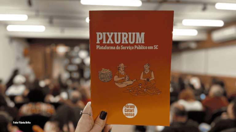 Em primeiro plano, uma mão segura um livreto de capa laranja, com o título em letras brancas: “PIXURUM – Plataforma do Serviço Público em SC”. No centro da capa, há uma ilustração de duas pessoas trabalhando artesanalmente com fibras, ao lado de um cesto. Na parte inferior, aparece a identidade visual do Fórum Catarinense em Defesa do Serviço Público. Ao fundo, desfocadas, diversas pessoas estão sentadas em um auditório