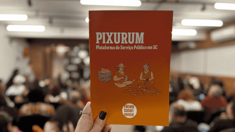 Em primeiro plano, uma mão segura um livreto de capa laranja, com o título em letras brancas: “PIXURUM – Plataforma do Serviço Público em SC”. No centro da capa, há uma ilustração de duas pessoas trabalhando artesanalmente com fibras, ao lado de um cesto. Na parte inferior, aparece a identidade visual do Fórum Catarinense em Defesa do Serviço Público. Ao fundo, desfocadas, diversas pessoas estão sentadas em um auditório