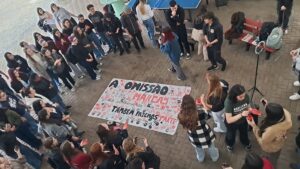 Fotografia feita de um ponto elevado mostra ato com servidores e estudantes. As pessoas formam uma roda ao redor de uma faixa estendida no chão com os dizeres: “A omissão deixa marcas. Quando escolhemos não agir, também fazemos parte.”