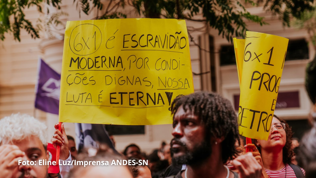 Pessoas em manifestação com cartazes. O cartaz principal tem escrito: "6x1 é escravidão moderna, por condições dignas, nossa luta é eterna!
